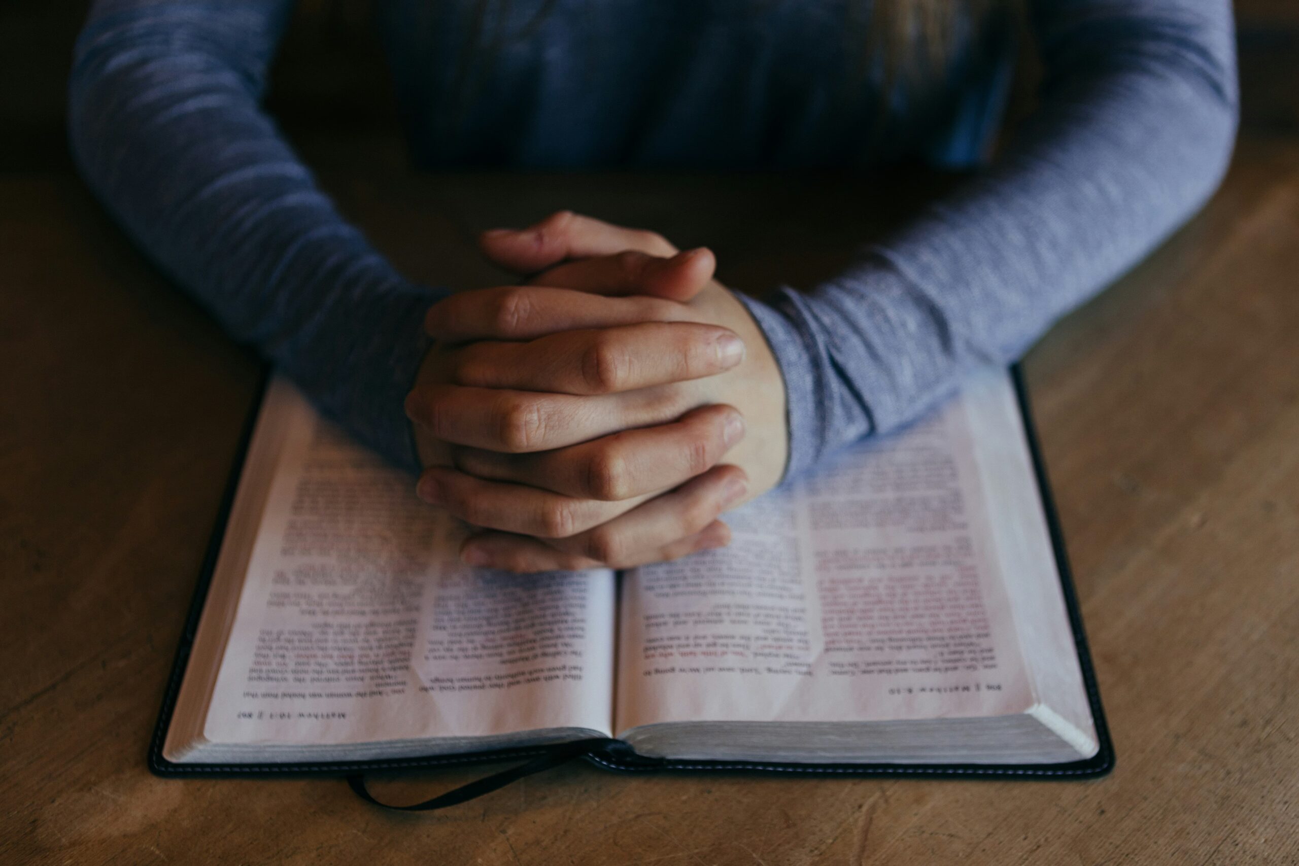 Person in a blue long sleeve shirt with clasped hands praying over an open bible
