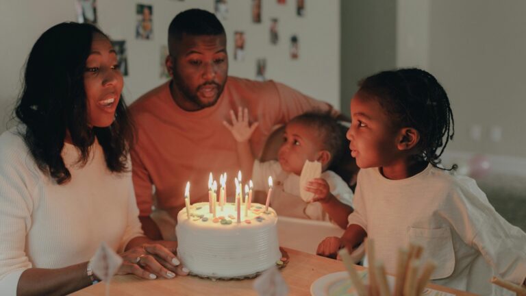 a mother, father, baby, and toddler gathered around a birthday cake on a table.