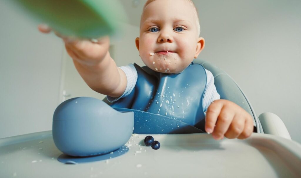 A baby wearing a bib and with a messy face holds out a plastic plate.