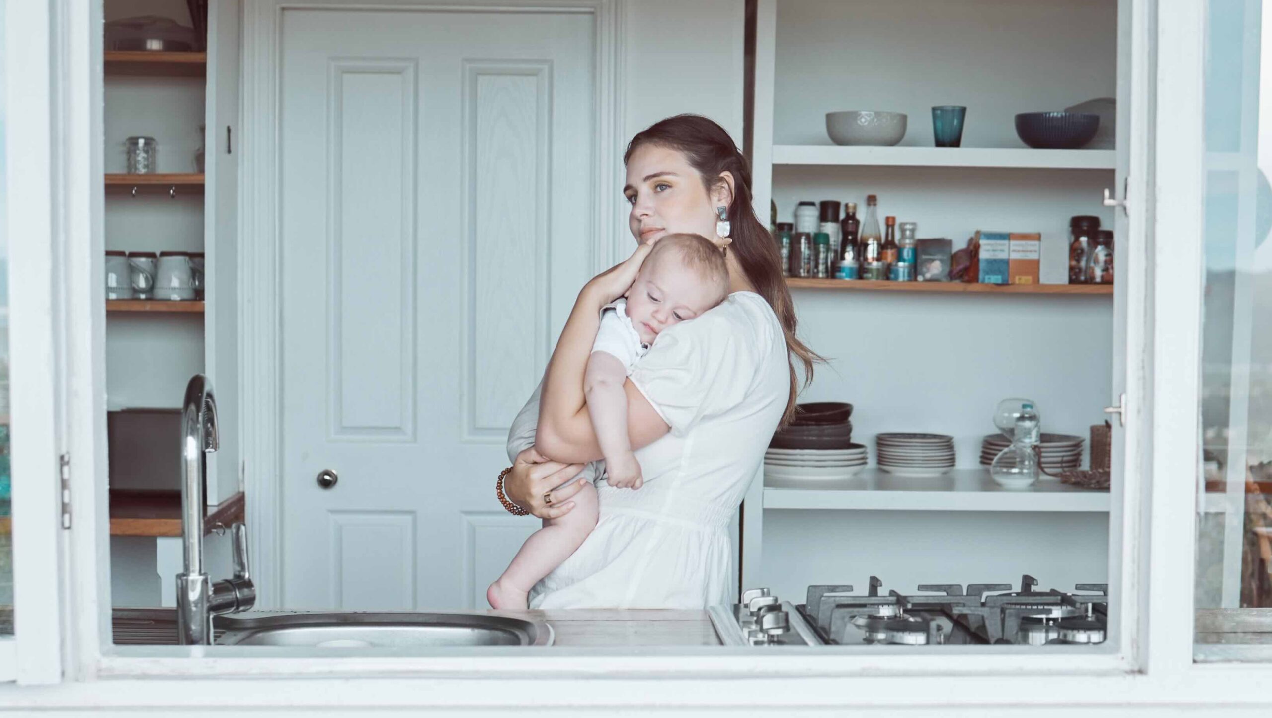 A woman holds a baby in a kitchen, calmly staring out an open window.