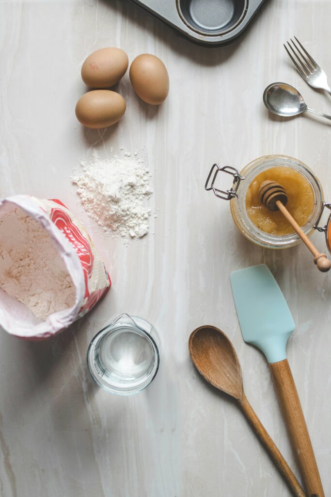 Overhead view of baking ingredients and utensils on a marble surface, including eggs, flour, honey, water, a spatula, wooden spoons, a fork, a spoon, and a muffin tin.