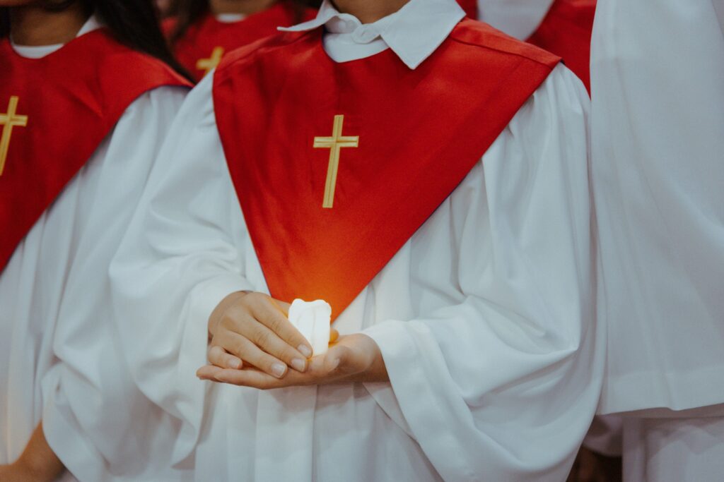 Child in a white robe with a red stole embroidered with a gold cross, holding a small lit candle during a religious ceremony.