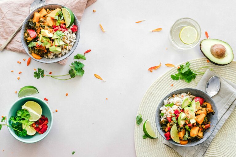 Overhead view of two colorful rice bowls with sweet potatoes, greens, avocado, red peppers, cilantro, and lime wedges, arranged on a light tabletop with extra herbs, lime, avocado, and a glass of water.