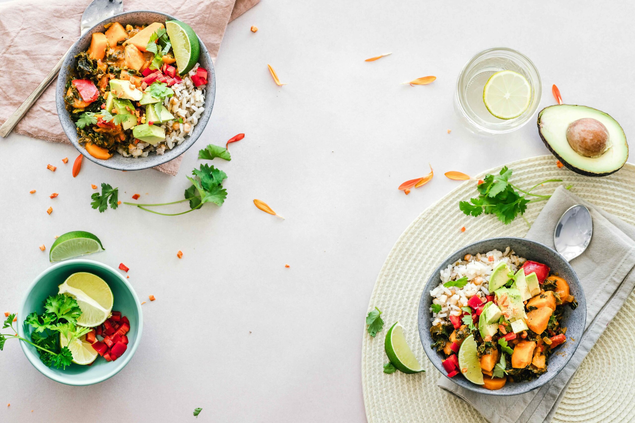 Overhead view of two colorful rice bowls with sweet potatoes, greens, avocado, red peppers, cilantro, and lime wedges, arranged on a light tabletop with extra herbs, lime, avocado, and a glass of water.