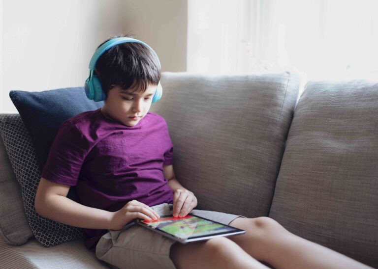A boy sits on a couch with headphones on while he plays on a tablet.