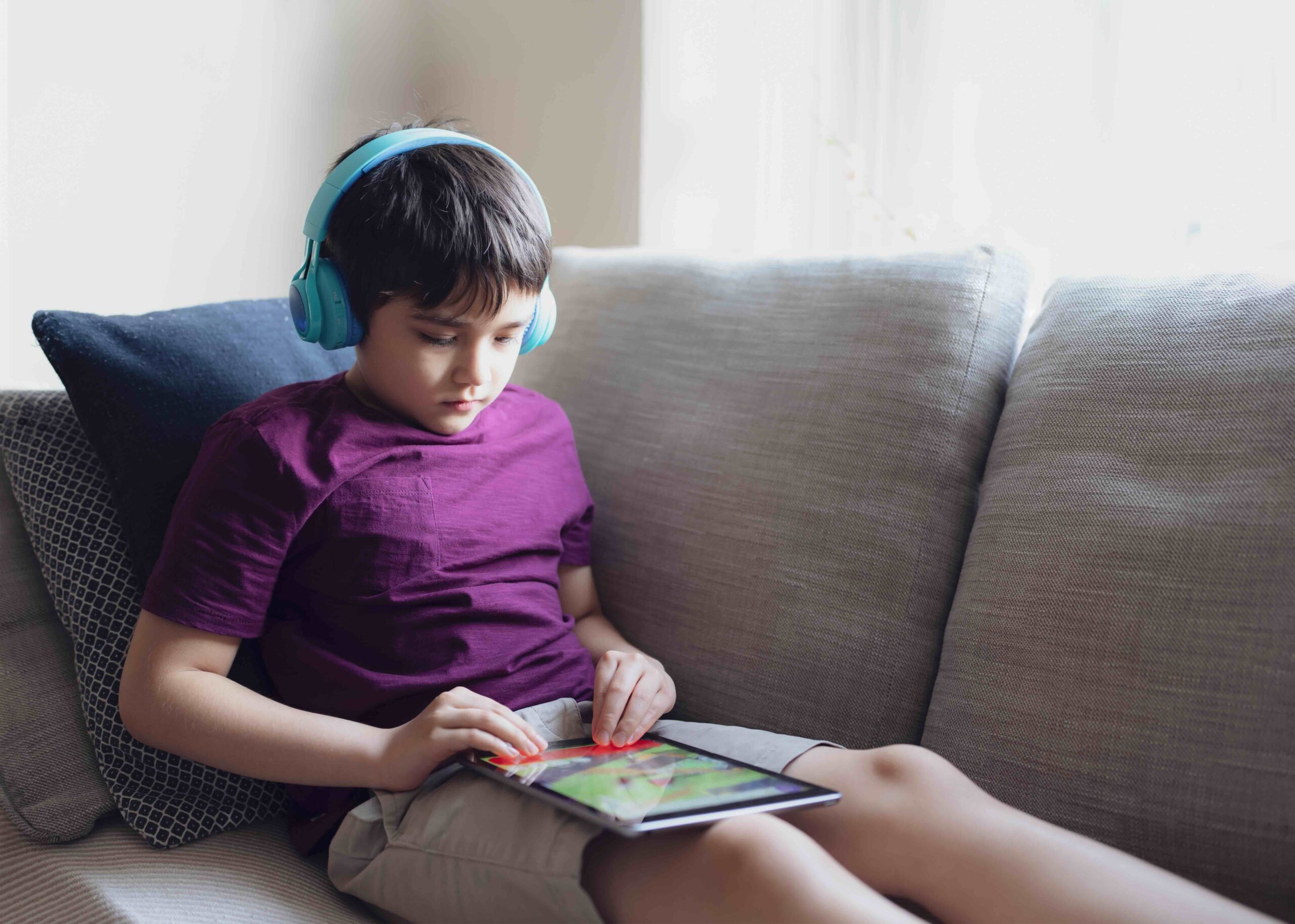 A boy sits on a couch with headphones on while he plays on a tablet.