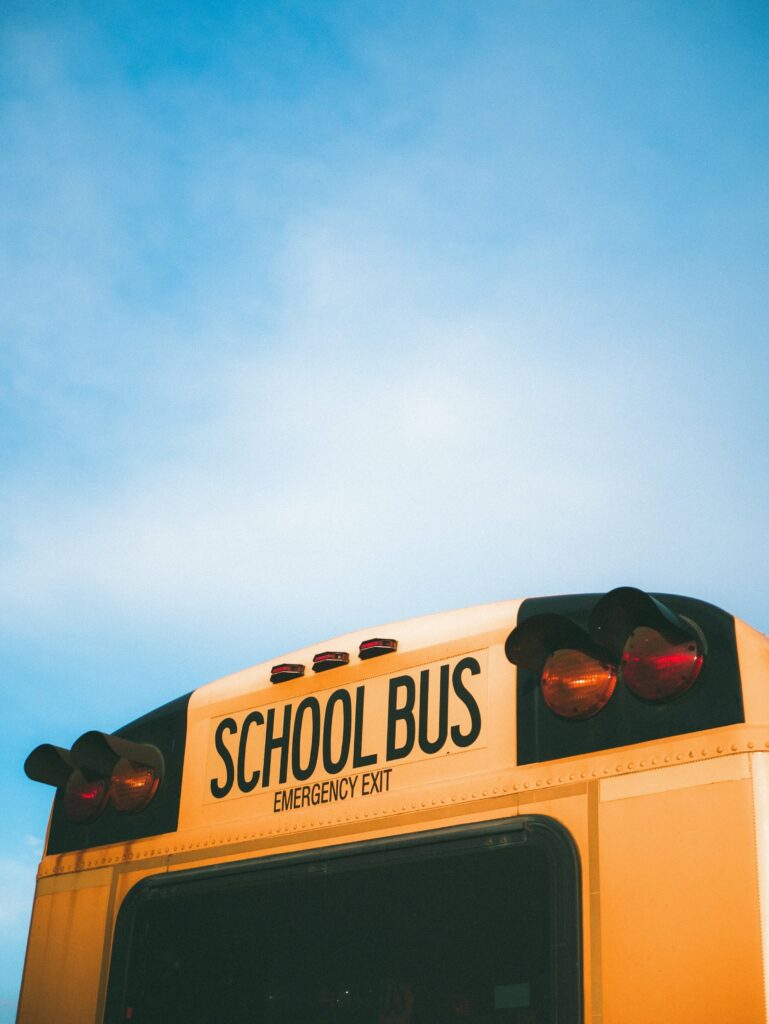 Yellow school bus viewed from the rear, with the “School Bus” and “Emergency Exit” signs visible against a bright blue sky.