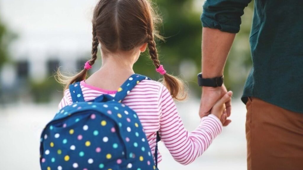 A child wearing a backpack holds her parent's hand as she makes her way to school.
